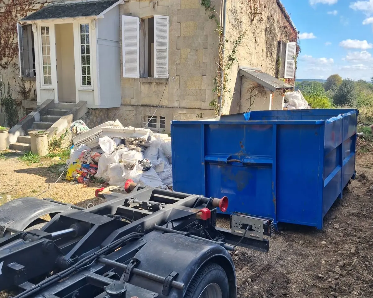 Benne ampliroll bleue installée sur un chantier de rénovation, à côté d’un tas de gravats et de sacs de déchets, le bras porte-benne au premier plan