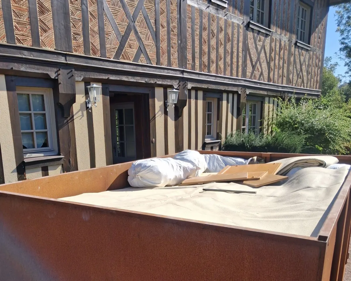 Benne chargée d’un matelas, d’un tapis roulé, de sacs et de planches de bois, devant une maison normande à pans de bois