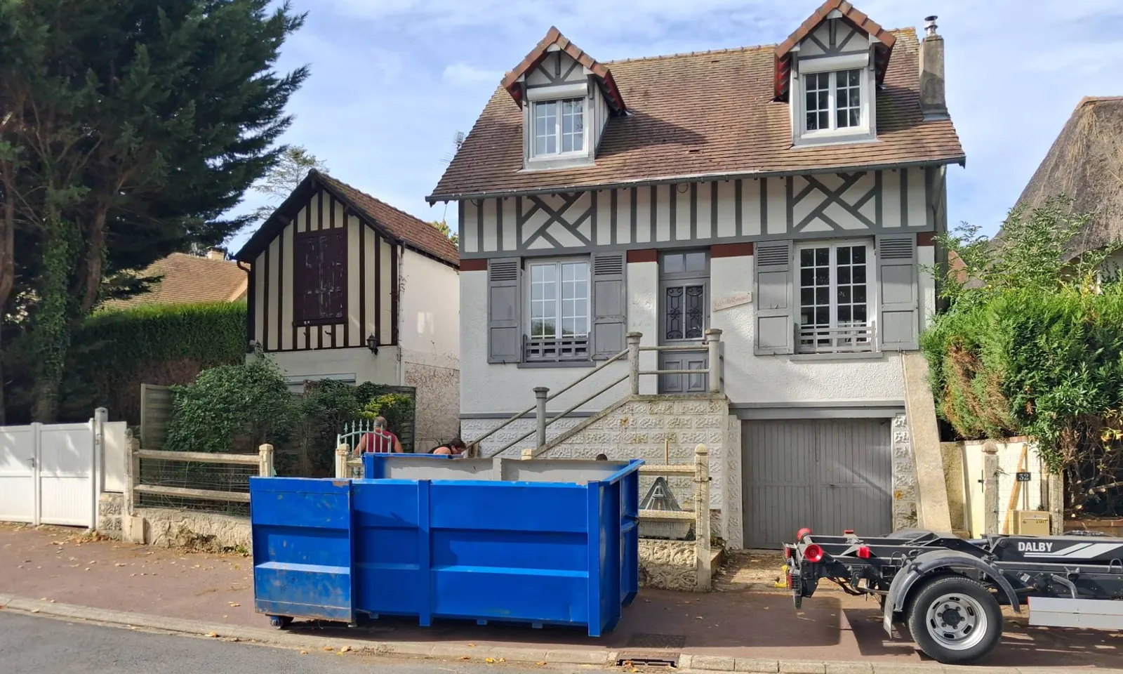 Benne ampliroll bleue posée en bord de voie devant une maison normande à colombages, la remorque porte-benne stationnée à droite