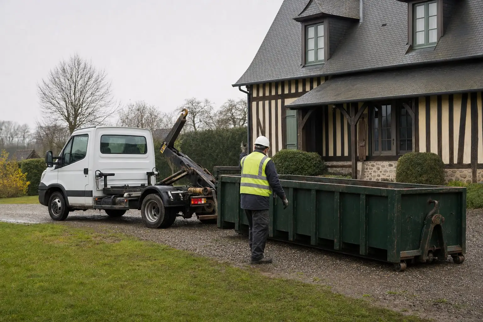 Porte-benne léger blanc déposant une benne vert foncé sur l’allée gravillonnée d’une maison normande, par temps couvert